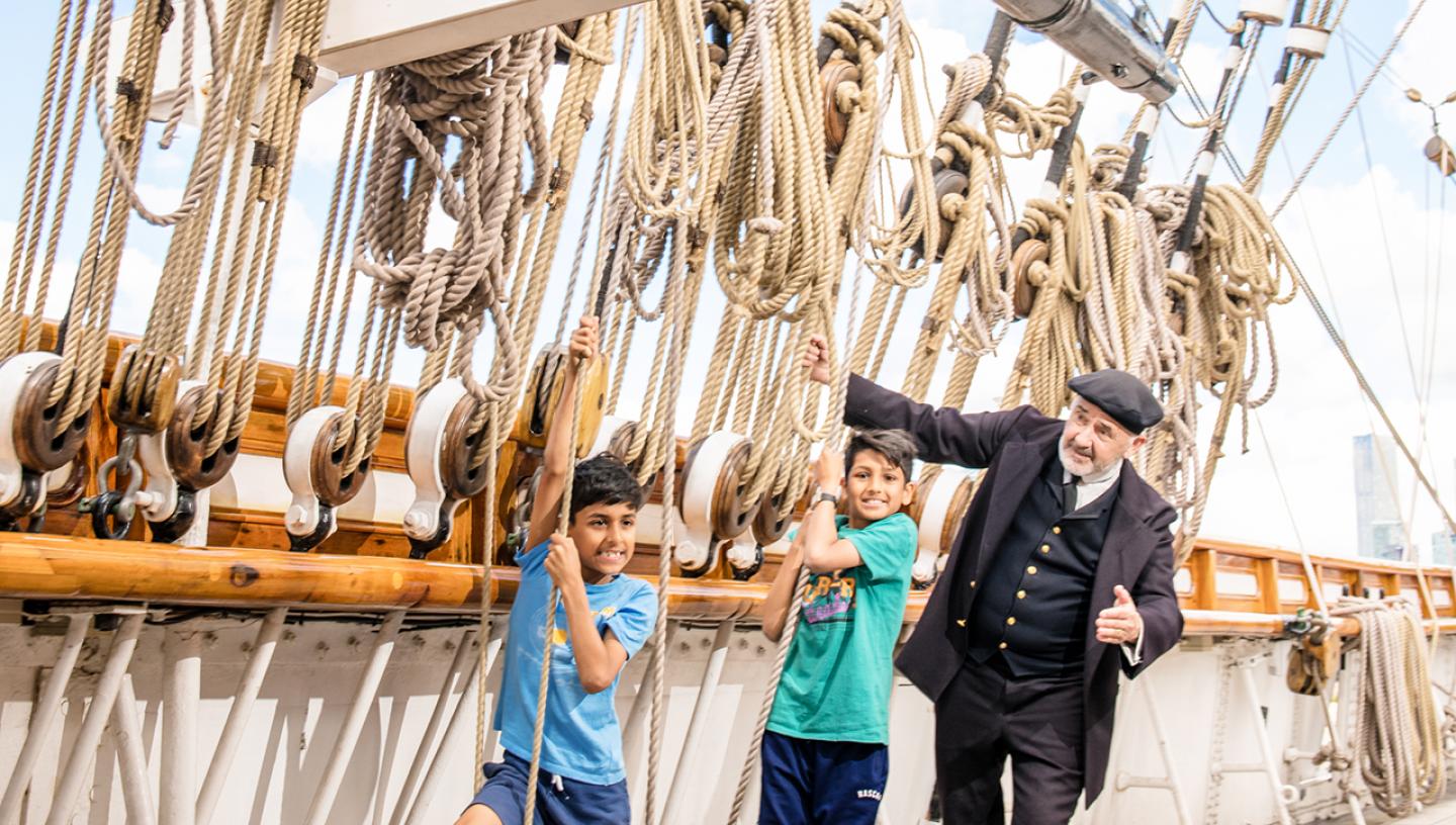 Two children and a character actor dressed as Captain Woodget play on the ropes of Cutty Sark. They are on the main deck, with a mass of ropes leading up to the rigging of the historic ship