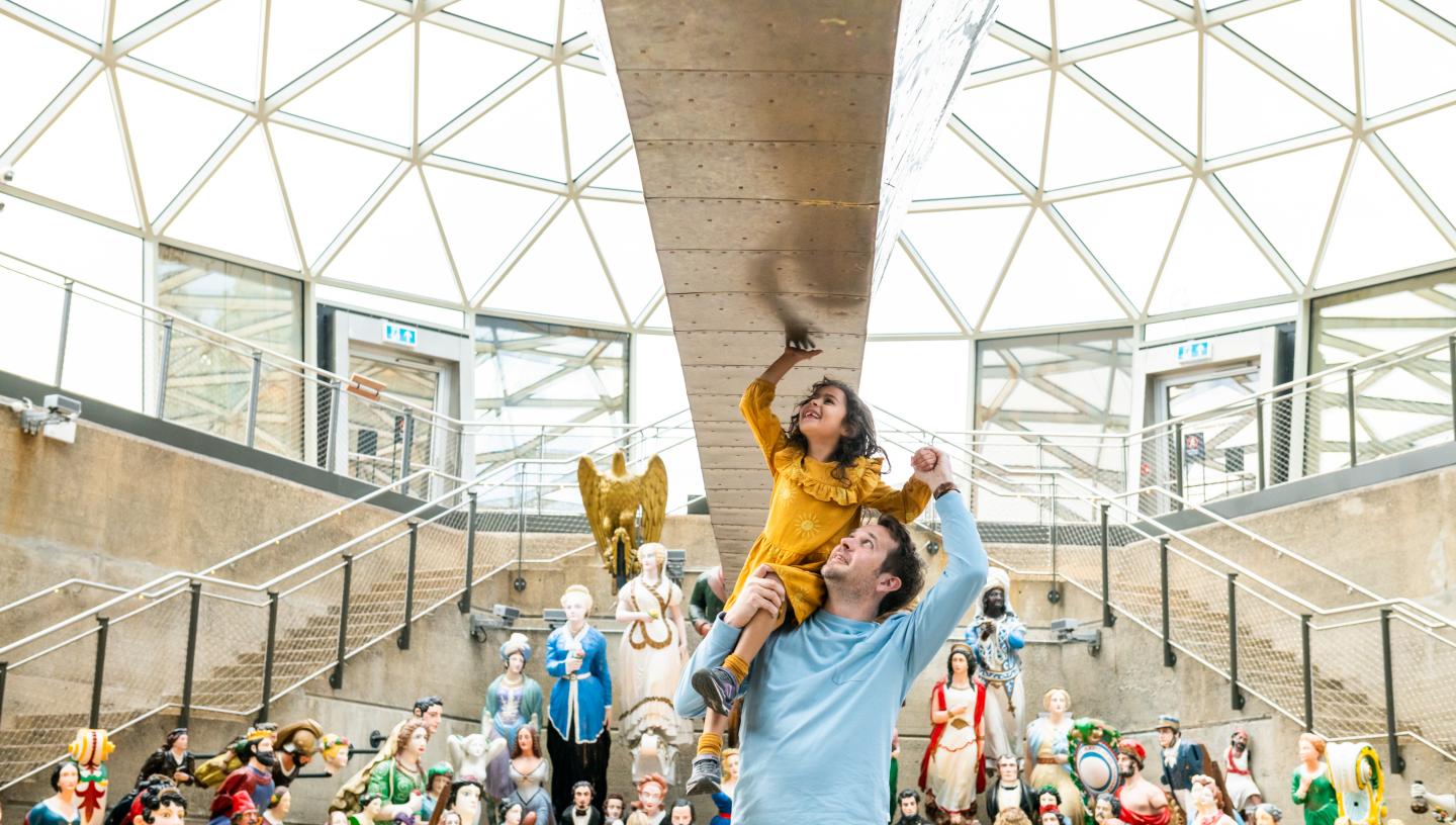 A father lifts his daughter up to touch the hull of historic ship Cutty Sark. A colourful display of ships' figureheads is arrayed in the background