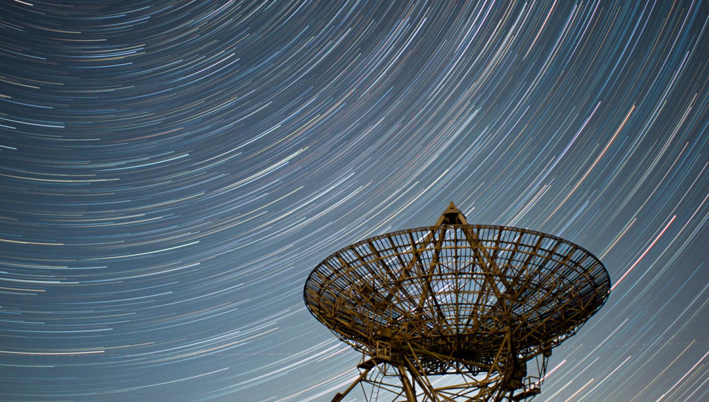 Image of radio telescope in the foreground and in the night sky behind it are semicircular star trails 