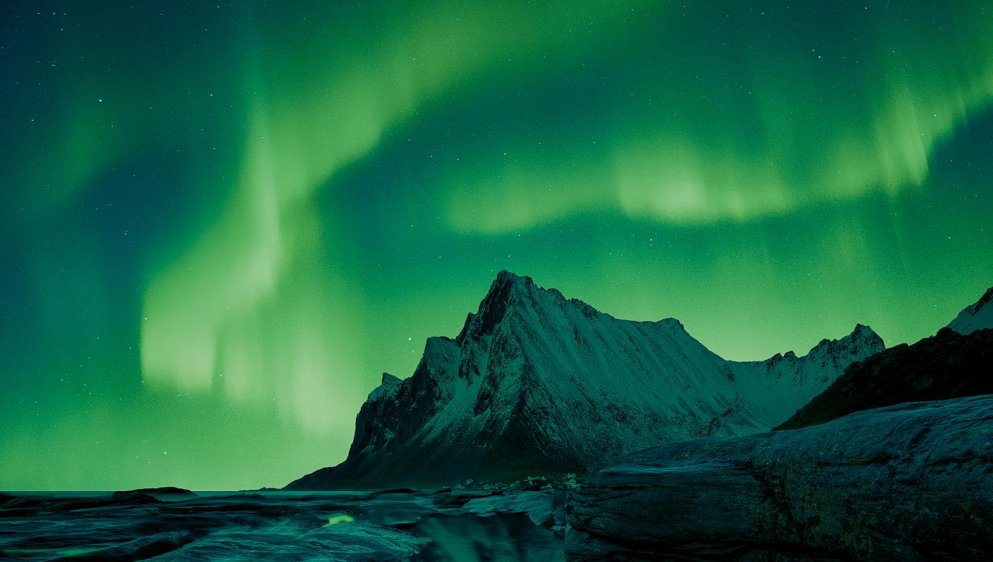 Image showing mountain and rocks in foreground, with rippling emerald aurora in the sky behind the mountain, which is reflected in a pool of water