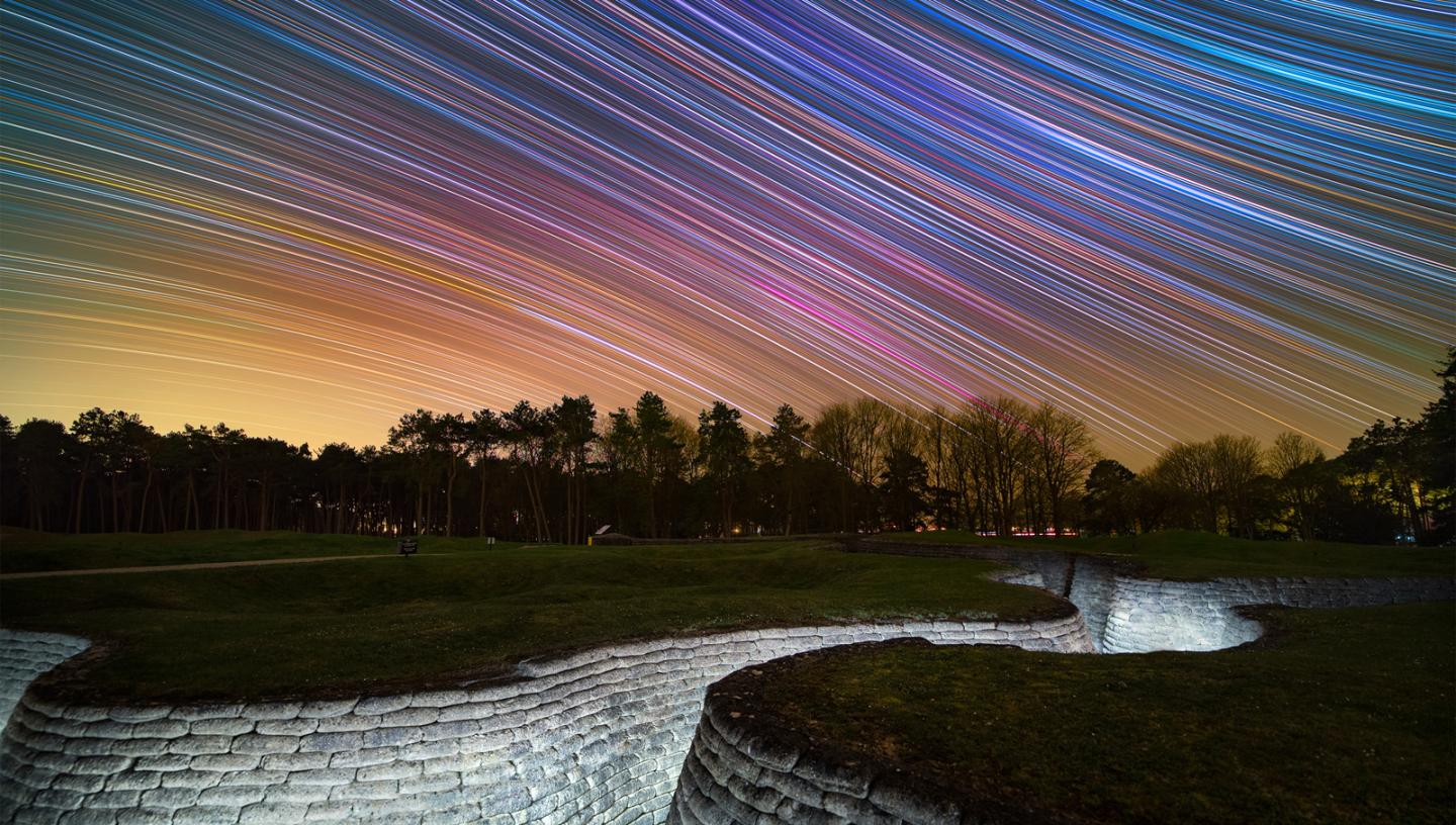 Image showing WWI trenches at bottom lit up with lights, a line of trees in the distance, and a purple, blue and yellow sky behind with star trails making curved diagonal lines