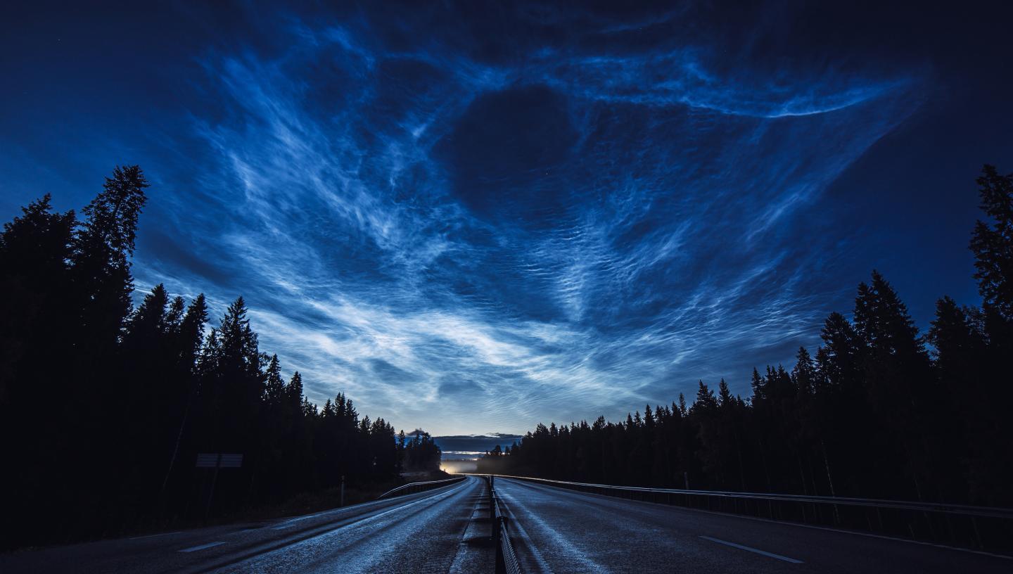 Image showing road at night with trees to the left and right, with sky above lit up with blue and silver noctilucent clouds
