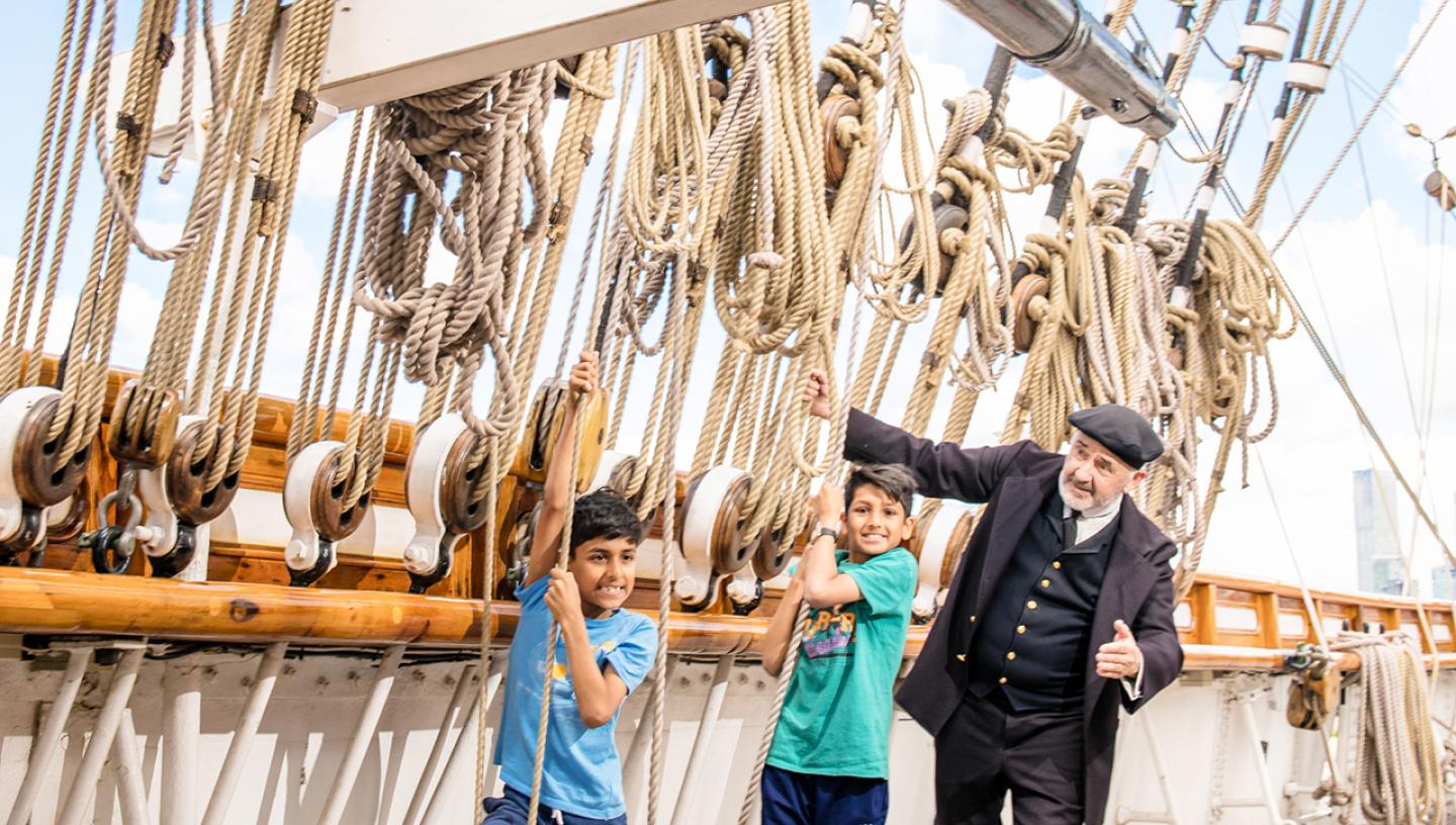 Two children pull on the ropes which are part of the tea clipper Cutty Sark's rigging, alongside captain Woodget
