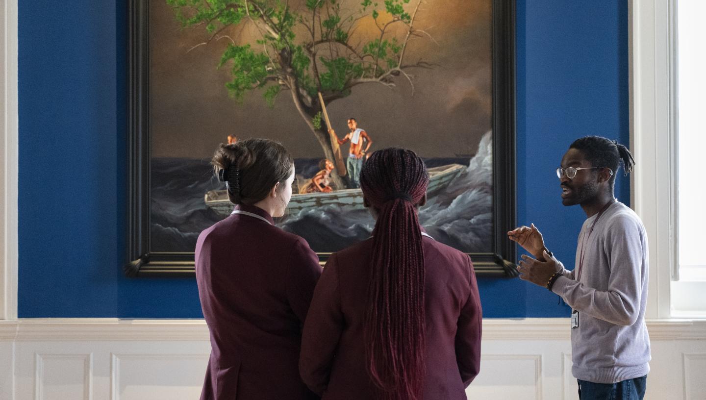 A group of school students in dark red blazers listen to an adult as he gives a talk in front of a painting