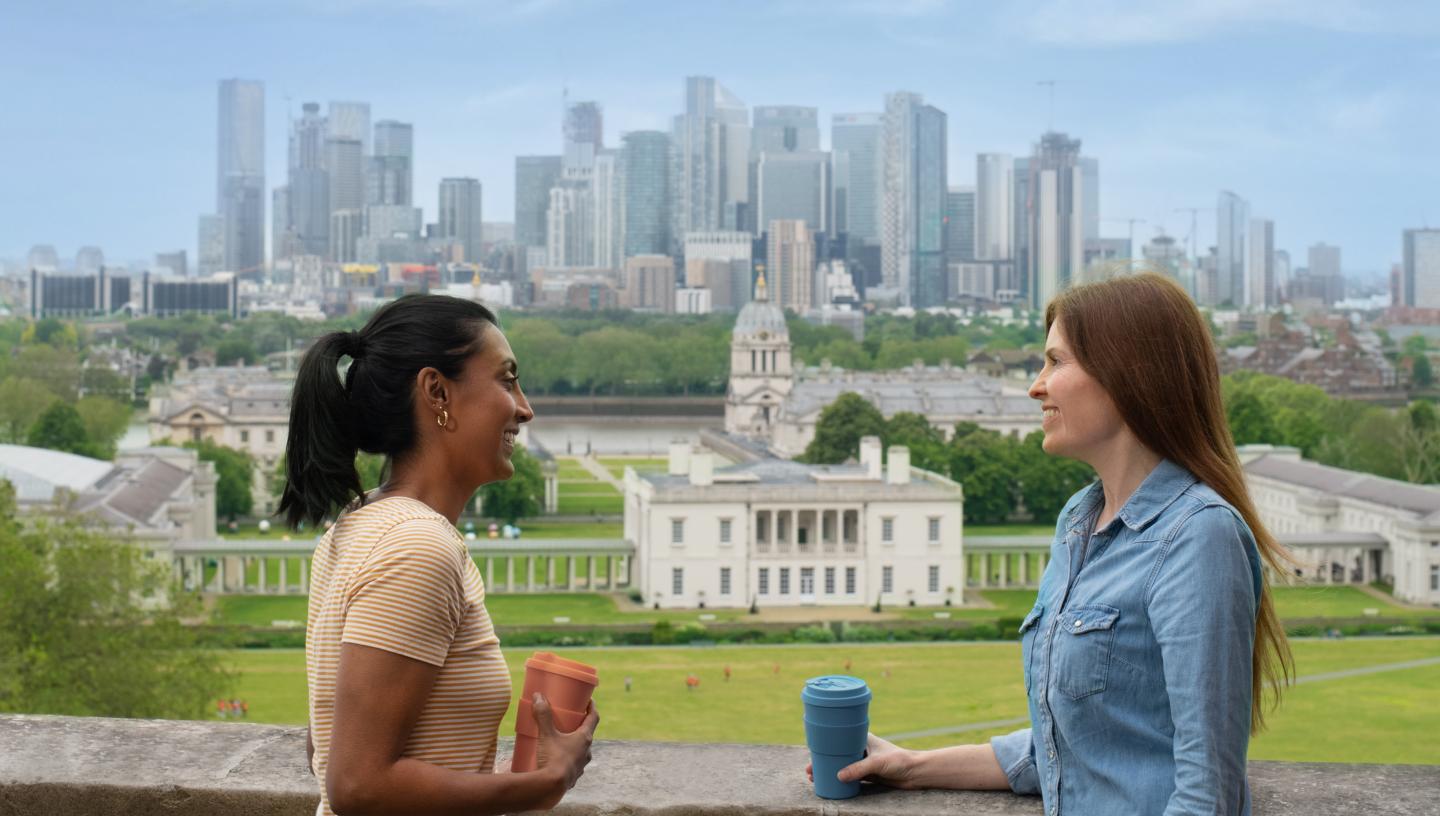 Two friends enjoy coffee with the view of Greenwich Park, the Queen's House and the skyline of Canary Wharf in the background