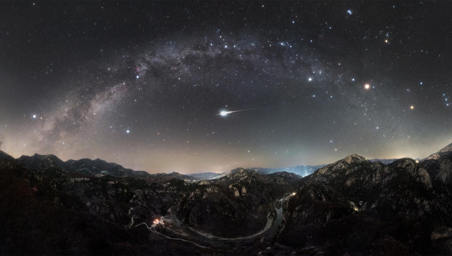 Rectangular image featuring a mountainous landscape in the bottom, with the Milky Way arching from the left to right of the image, smattered with stars. In the direct middle of the arc is an Orionid meteor, shining very brightly
