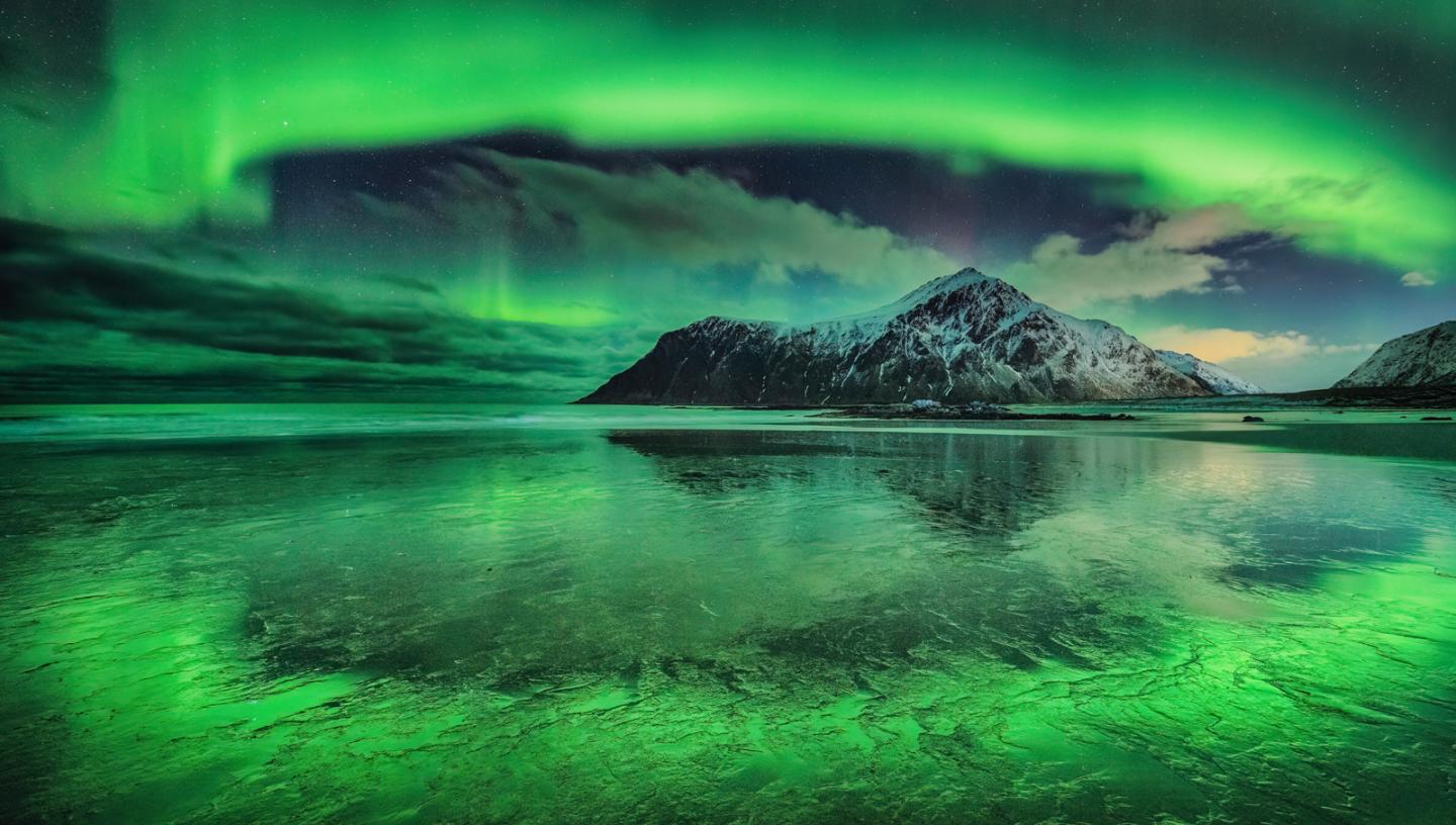 Image of a snow-capped mountain next to a wide expanse of frozen water, with a vivid aurora making almost an arc in the sky from left to right, which is reflected in the ice below