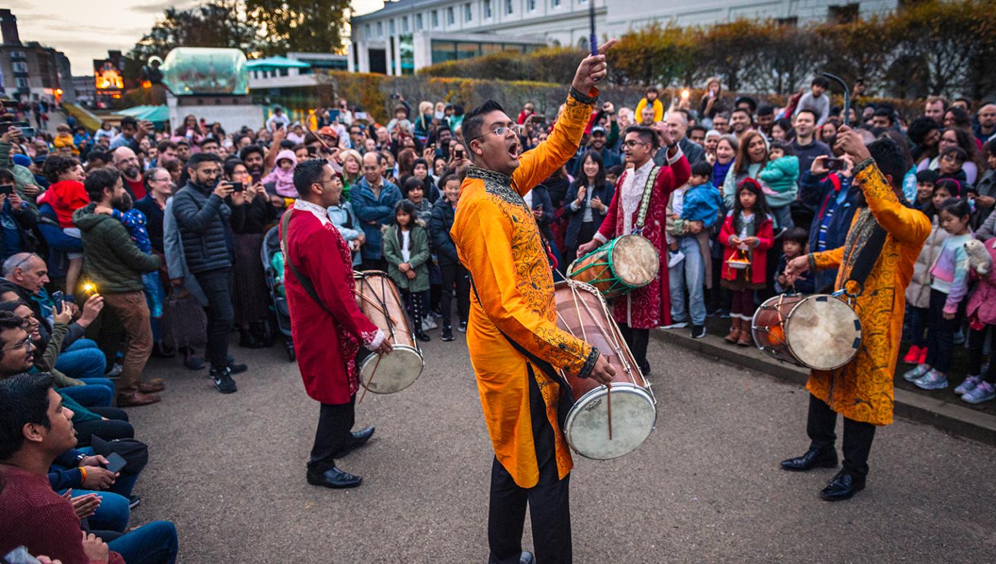 A group of south Asian drummers perform outside the National Maritime Museum in Greenwich at dusk, with a crowd looking on 