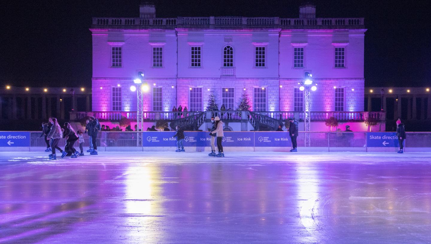 Image of the Queen's House Ice Rink with the classical building of the Queen's House in the background, lit up purple and blue, with a quite empty rink in front of it, only around 8 people on it