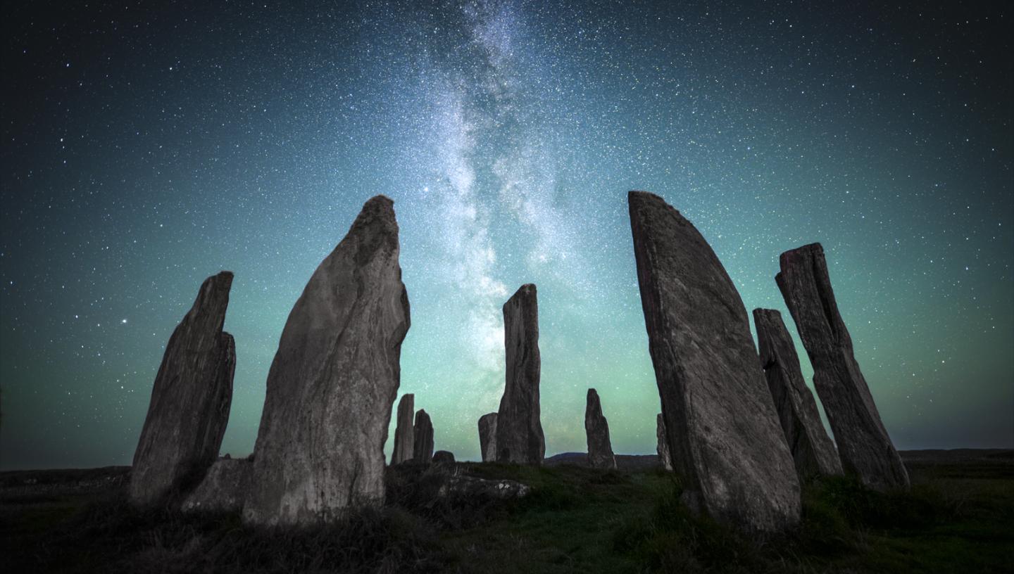 Image of a prehistoric rock formation akin to Stonehenge in the foreground of the image, with the Milky Way rendered in blues and greens in the sky