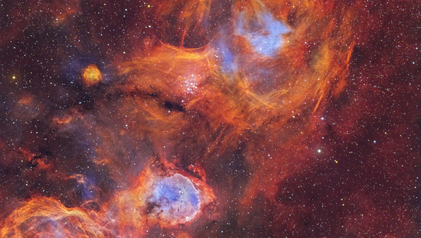 Image showing vivid nebulae in hot oranges and reds and cool blues, resembling a flame. The background is a starry sky coloured in dark reds with hundreds of stars