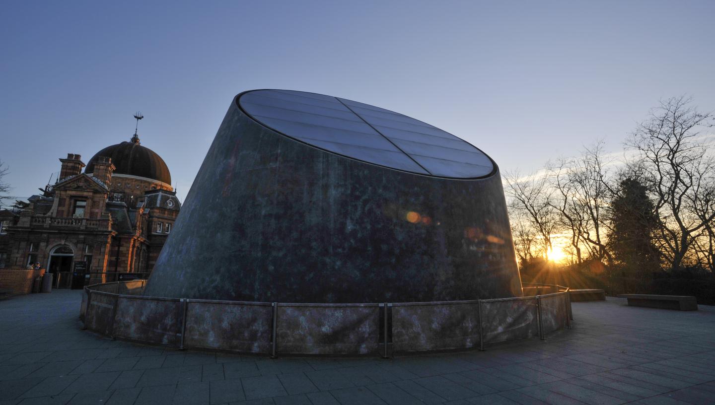 The grey metallic dome of the Peter Harrison Planetarium, with the historic buildings of the Royal Observatory Greenwich behind. A low sun is seen just peeping through the trees in the background