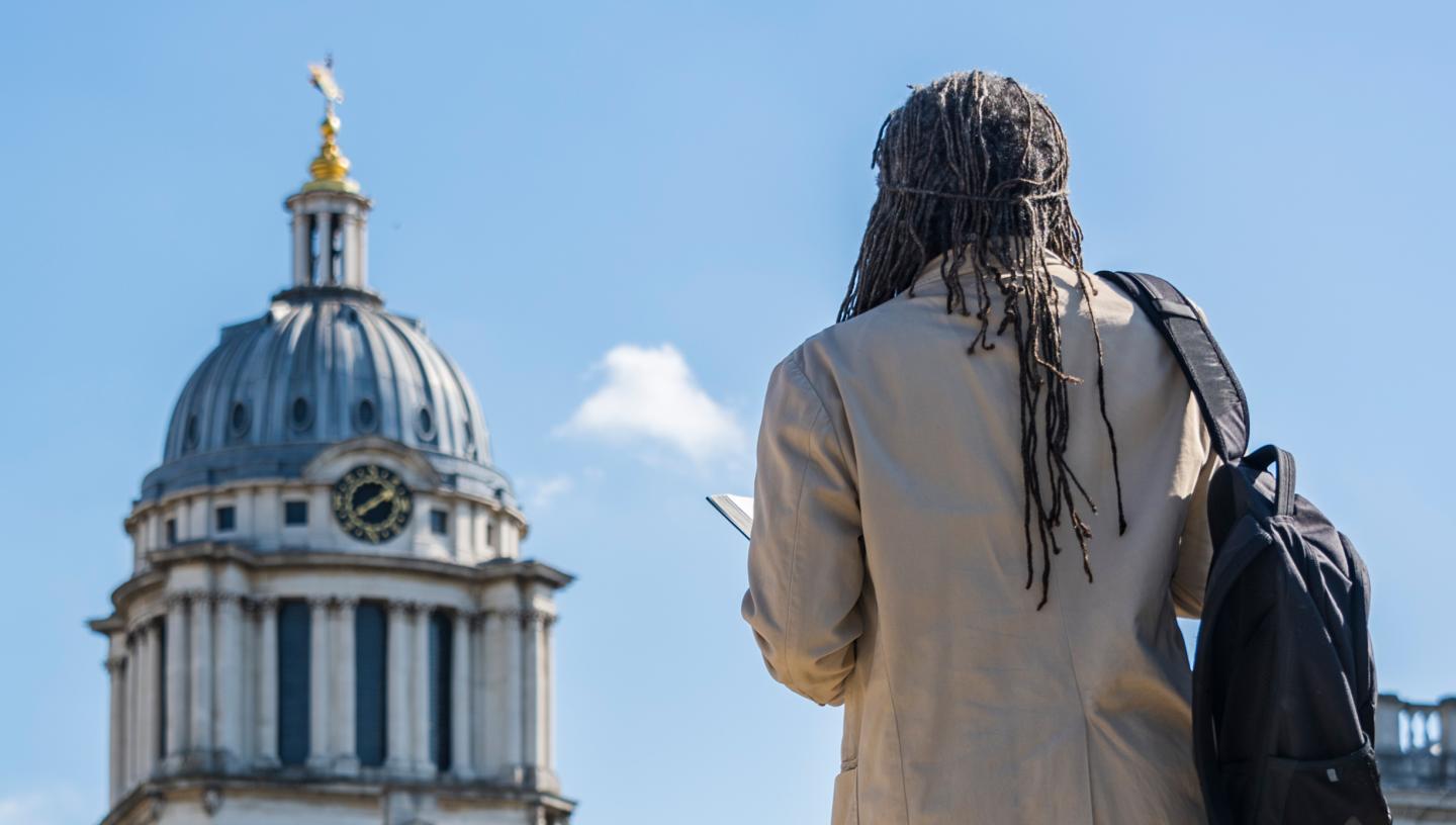A man stands with his back to camera, looking up at a large round clock tower, part of the Old Royal Naval College building in Greenwich