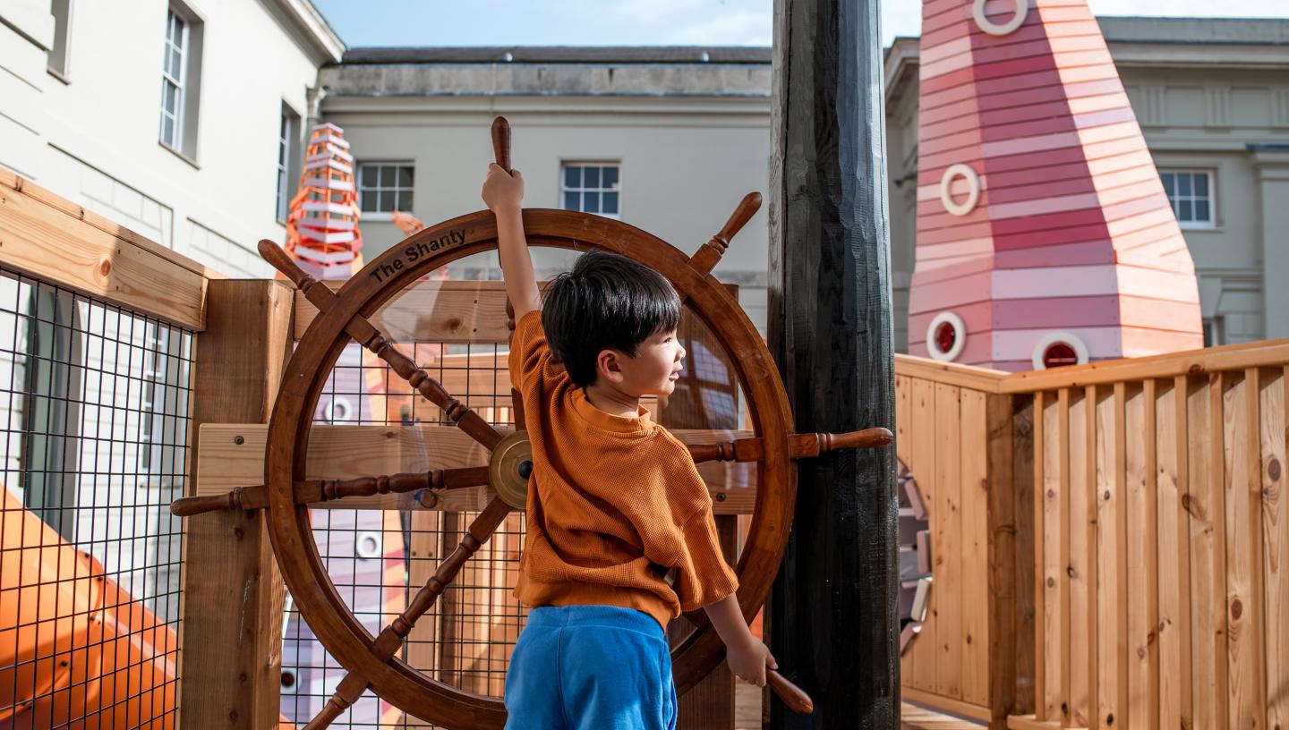 A young boy takes the helm of a large ship's wheel, part of an outdoor playground apparatus at the National Maritime Museum