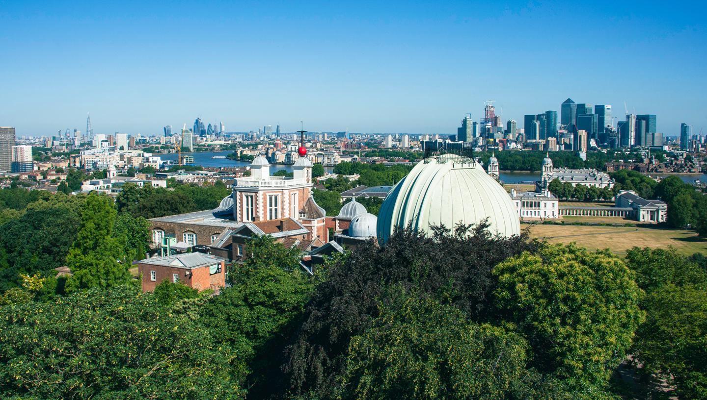 An aerial view of the Royal Observatory Greenwich, looking across the observatory domes and historic buildings towards the River Thames, with the London skyline in the distance