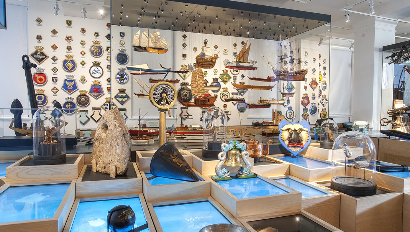A photo of the Sea Things gallery in the National Maritime Museum, with lots of maritime objects displayed in glass cases, wooden platforms and mounted on the walls