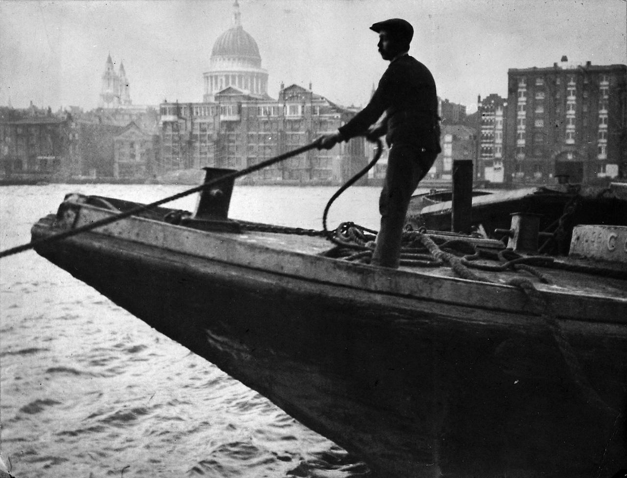 An image showing 'A lighterman standing on the bow of a lighter, hauling on a rope'