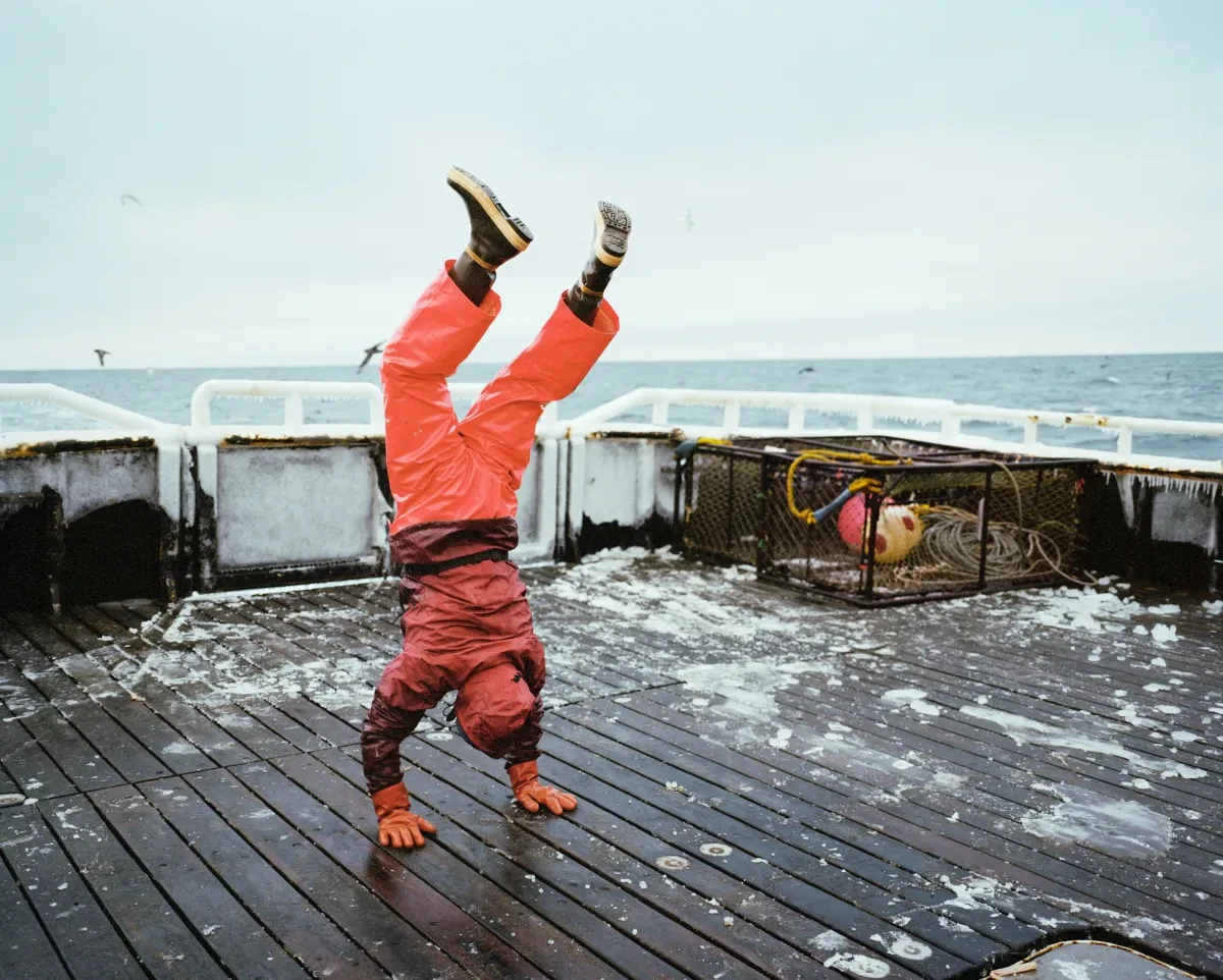 Image shows a man in an orange work suit doing a handstand on an icy deck