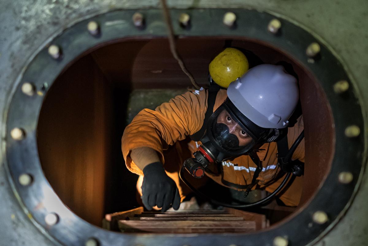 A man climbs down a hatch on board an oil tanker in heavy protection equipment, including a gas mask and visor
