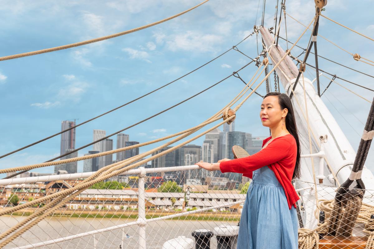 A woman in a red cardigan stands on the deck of Cutty Sark admiring the view towards Canary Wharf