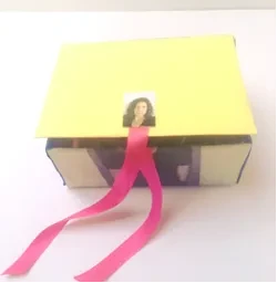 A yellow cardboard box with a pink ribbon and a photo on top