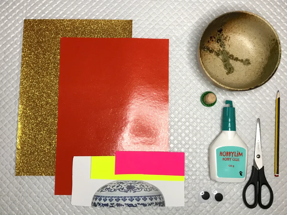 Materials for the activity: gold and red paper, small bits of coloured paper, glue, scissors, a bowl, a small lid, a pencil and two googgly eyes are optional.