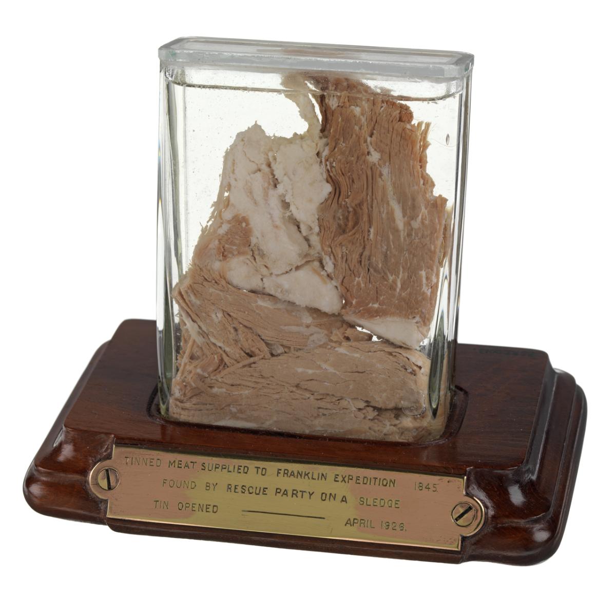 A clear glass display case containing a piece of preserved meat. The inscription reads 'TINNED MEAT SUPPLIED TO THE FRANKLIN EXPEDITION 1845. FOUND BY RESCUE PARTY ON A SLEDGE. TIN OPENED-APRIL 1926'