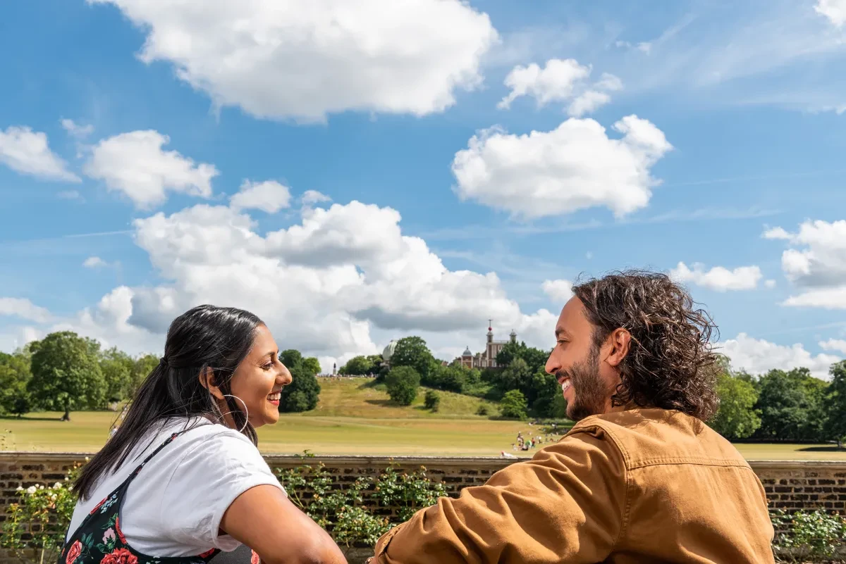 A couple sits on a bench with Greenwich Park and the Royal Observatory in the background