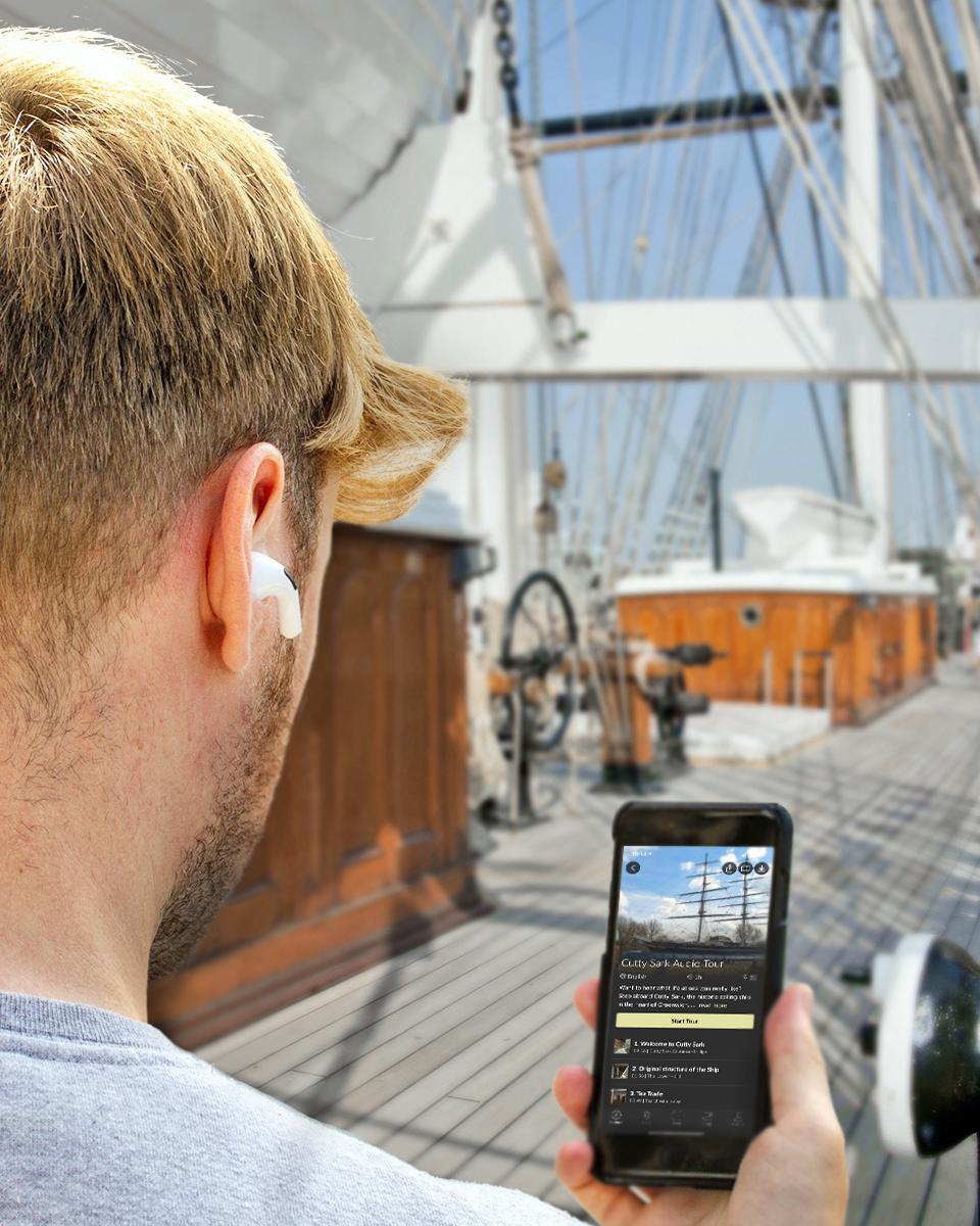 A man listens to the Cutty Sark audio guide on his phone, with the main deck of the ship in the background