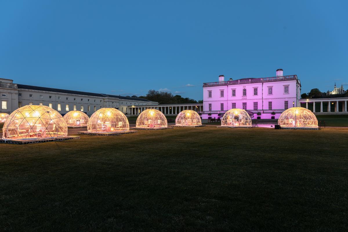 Outdoor dining pods lit up at night with the Queen's House in Greenwich, London behind