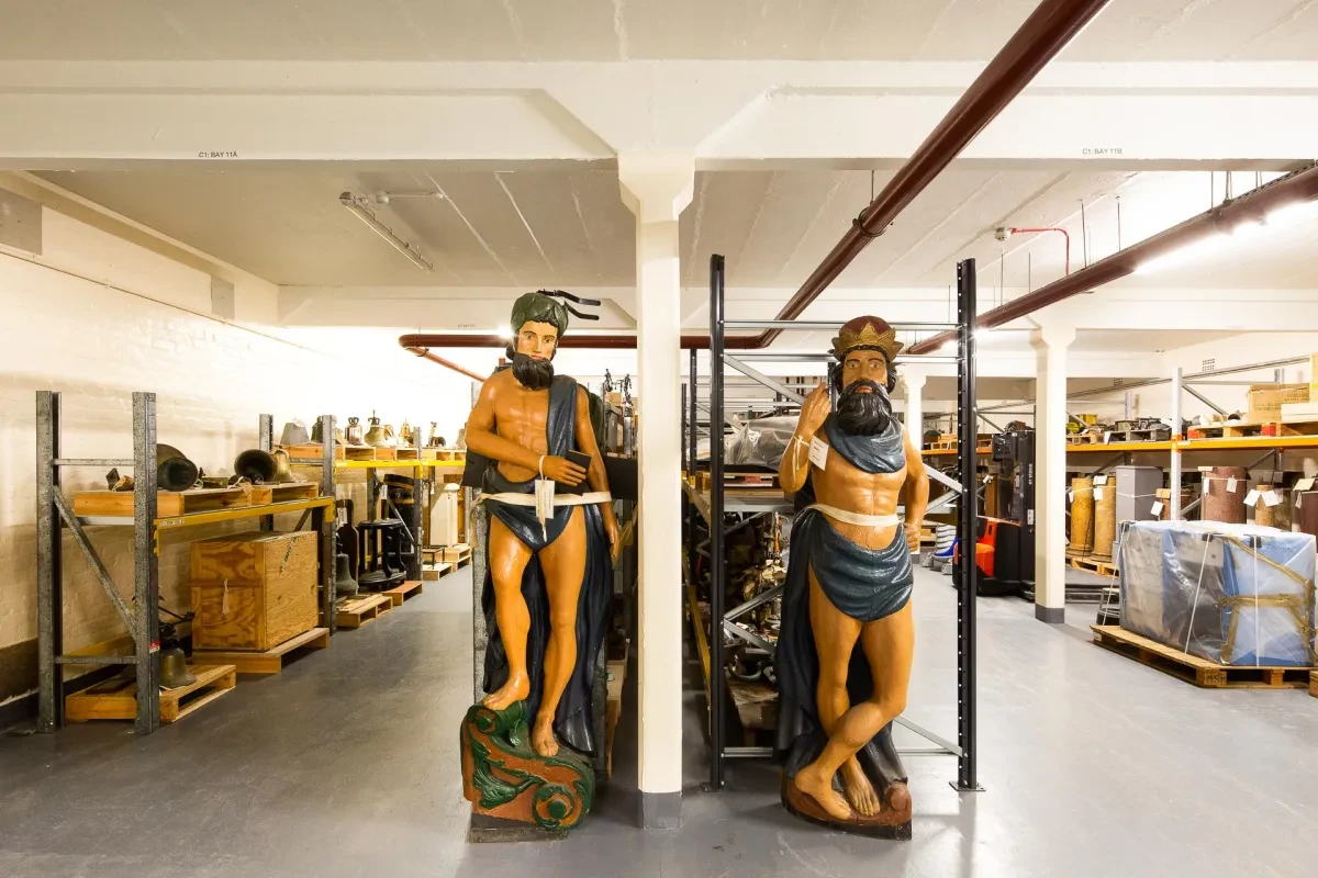 Inside Royal Museums Greenwich collections centre - rows of objects including two ships figureheads
