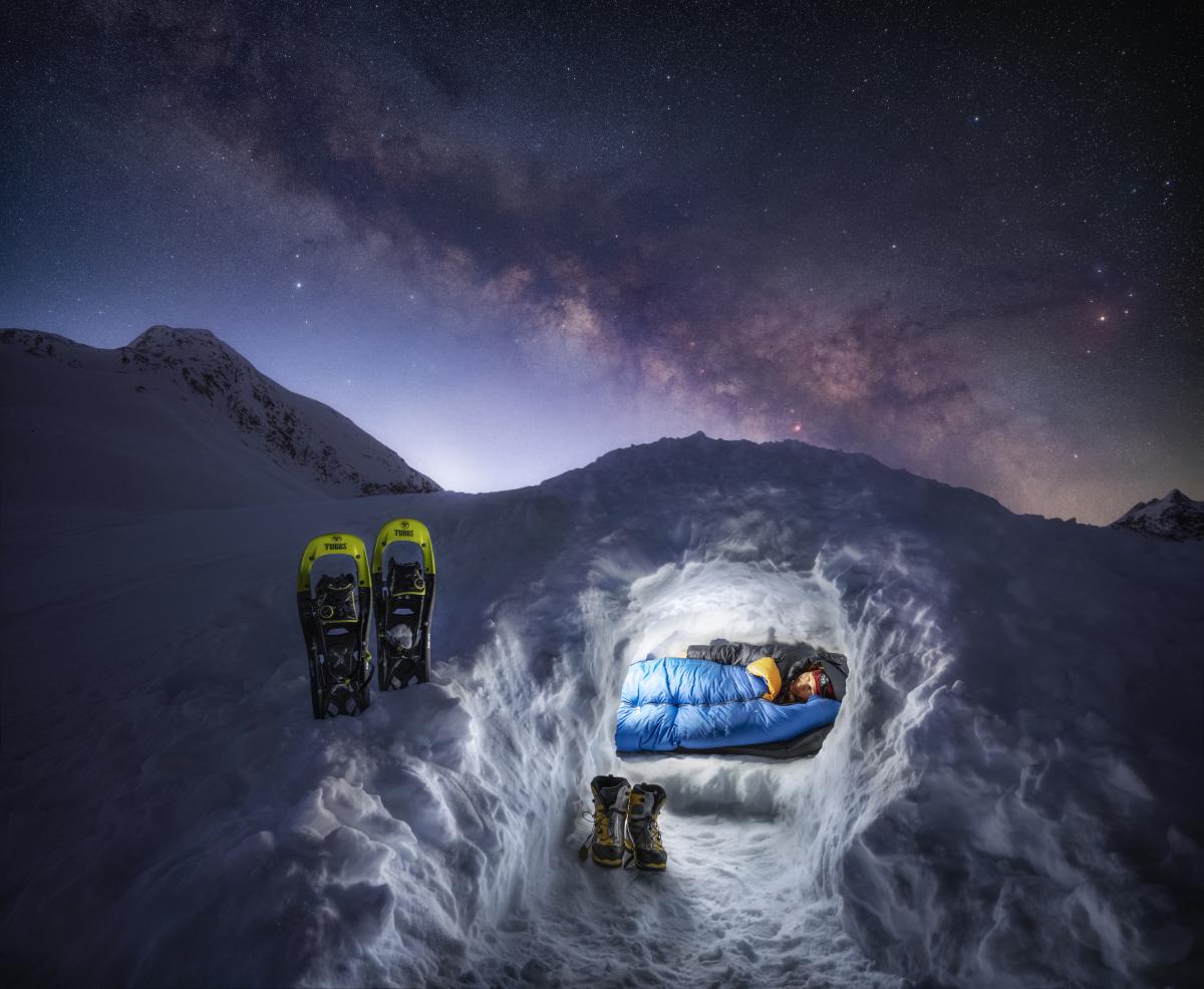 Man wrapped up in a sleeping bag in an igloo with shoes outside and the Milk Way in the sky
