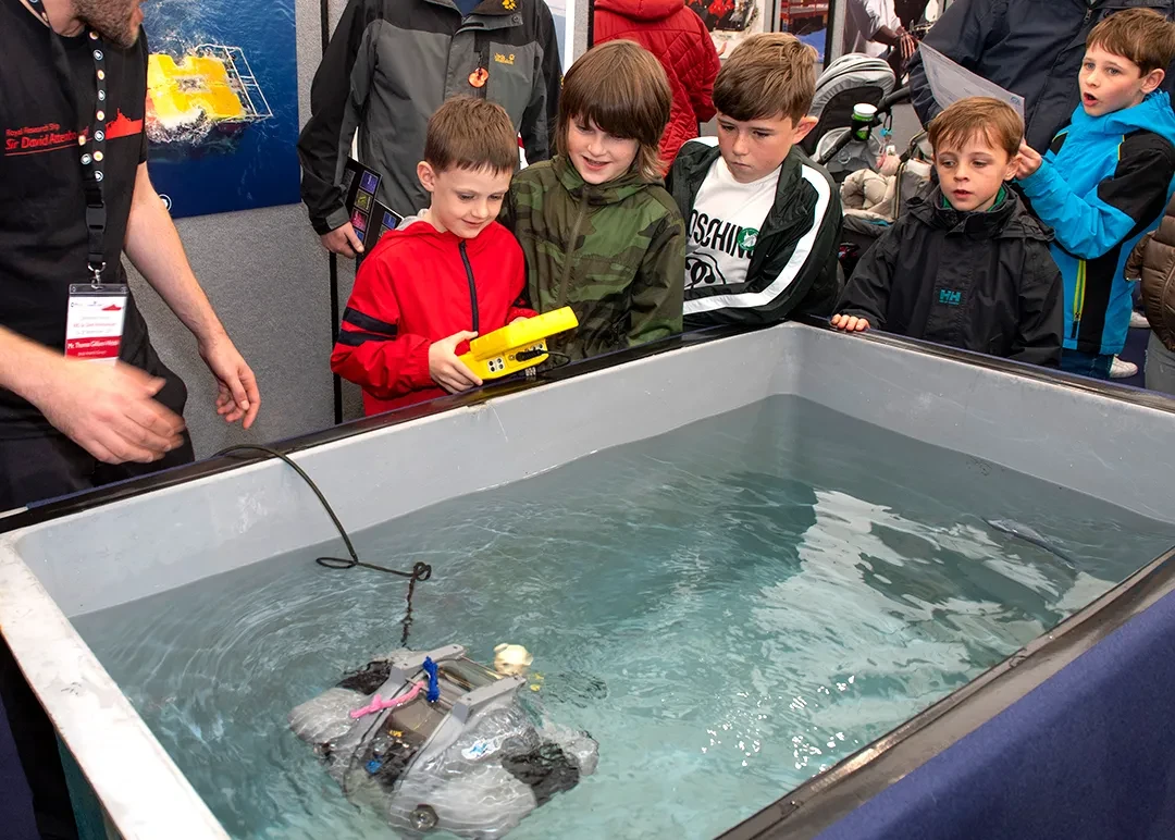 A boy controls a remote control submersible in a water tank as other children watch on