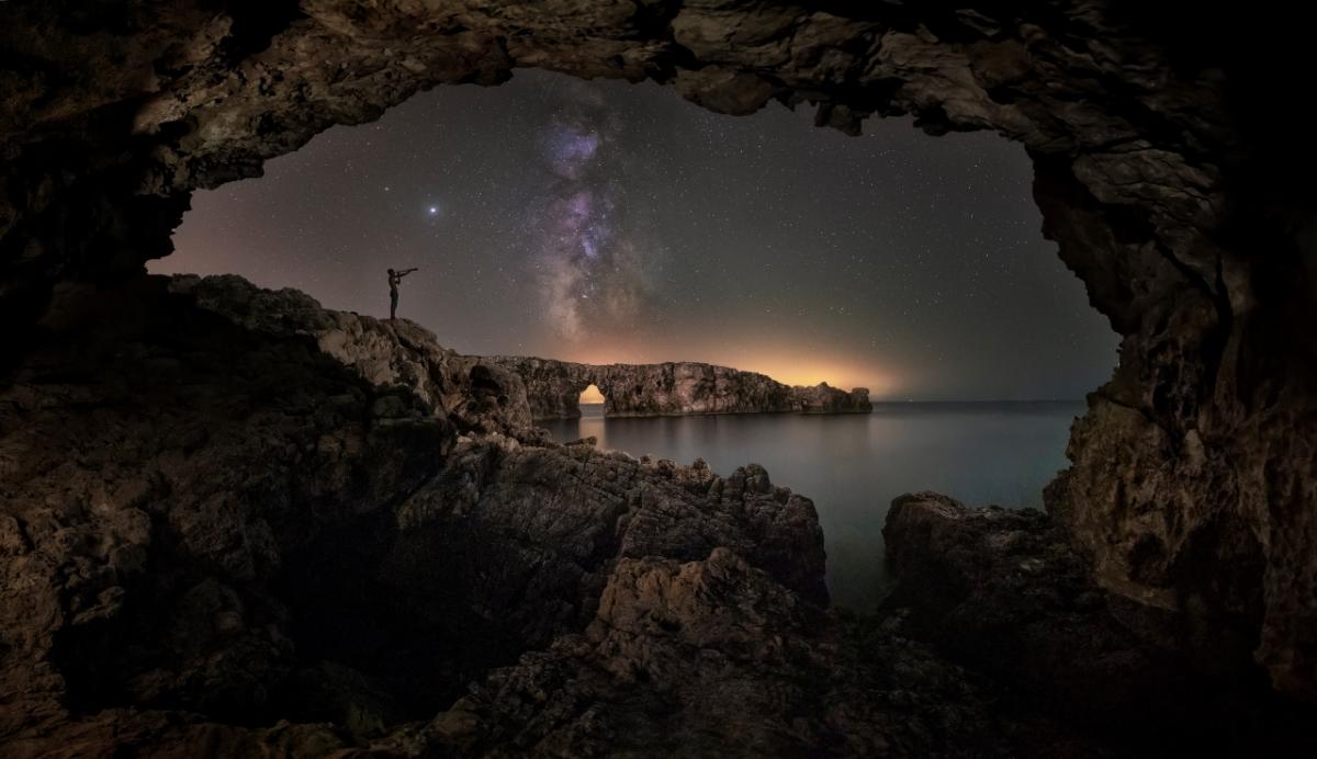 A silhouette of a person looking out to sea at night, with rocks and stars framing them