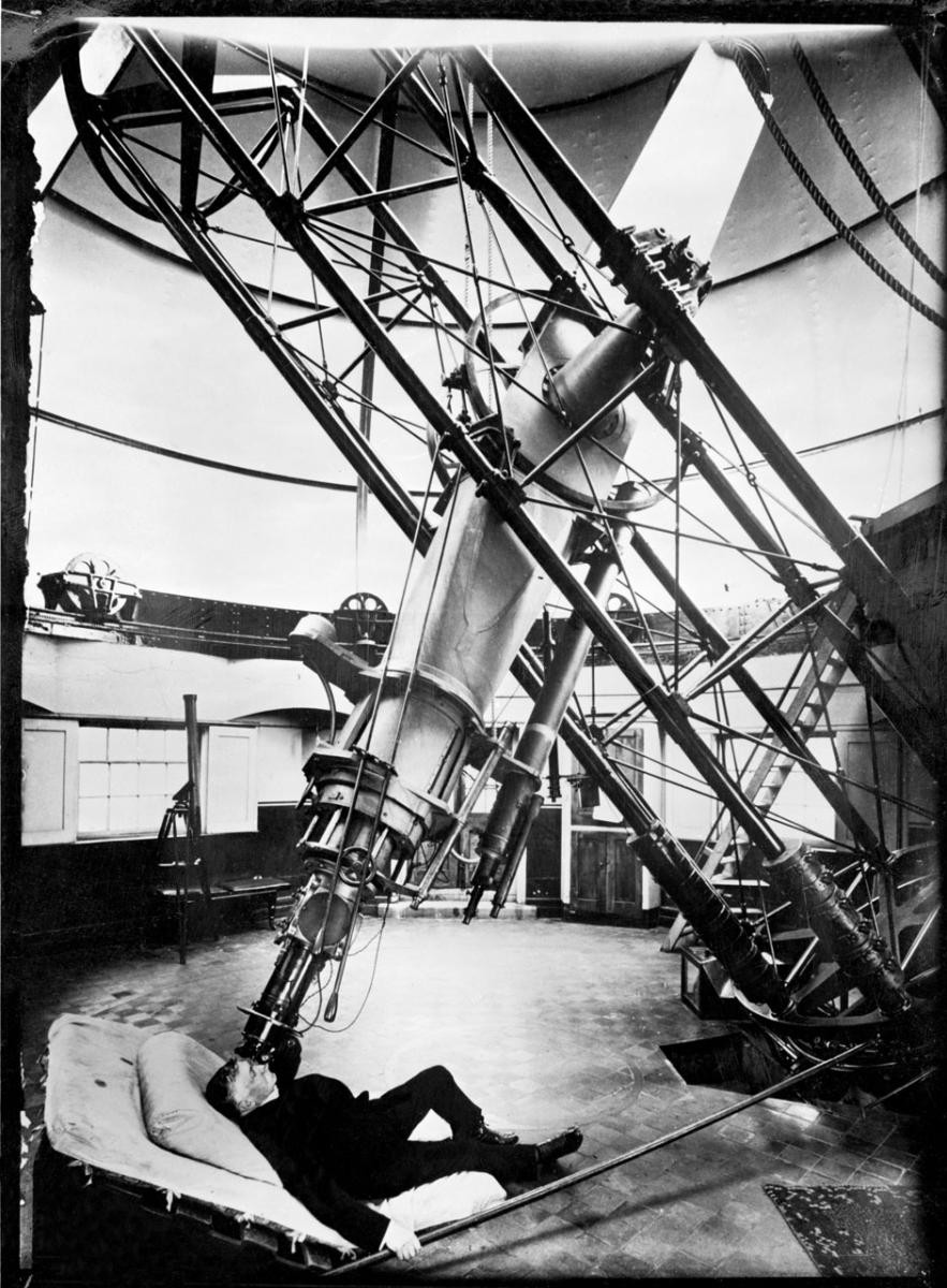 Historic black and white photograph of an astronomer using the Great Equatorial Telescope in Greenwich. He is lying on a reclining chair looking through the eyepiece, with the huge 28 feet-long telescope above him