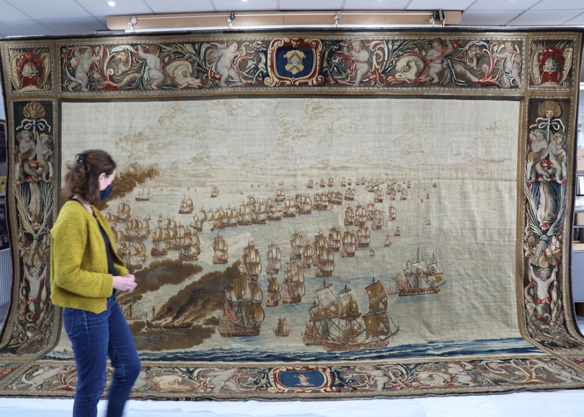 A woman walks in front of a large tapestry during conservation work. The tapestry shows a naval scene and is suspended from the ceiling, with part of the bottom border trailing on the floor
