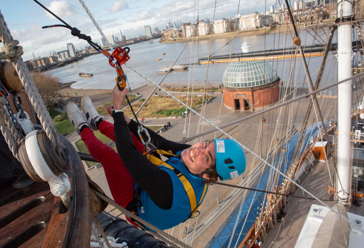 A man hangs from the rigging of Cutty Sark before taking a zip line back to street level. The River Thames is below