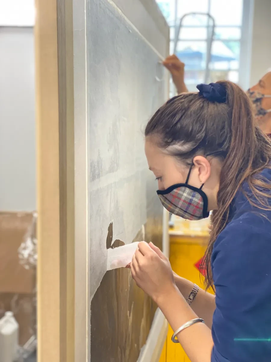 A painting conservator removes a layer of protective tissues from the front of a painting during conservation