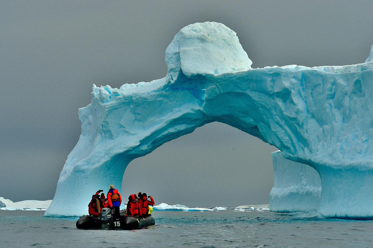 An ice arch in Antarctica with people looking on from a small motorboat