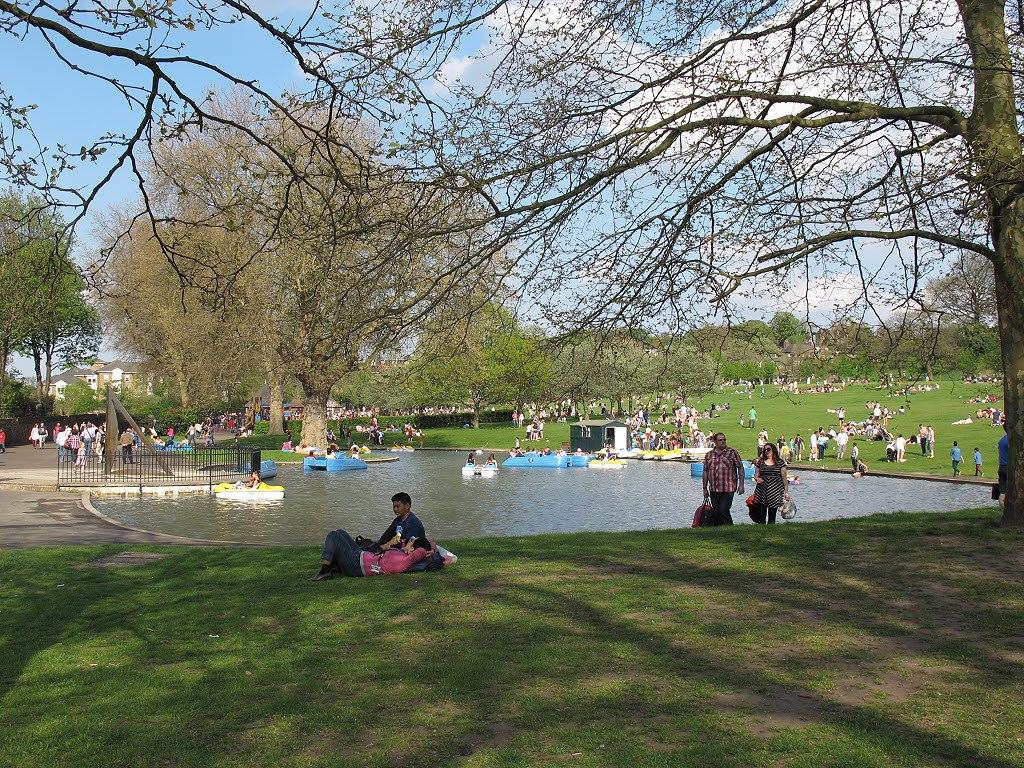 A photo of the boating lake in Greenwich Park, with people lying on the grass in the foreground and background, and blue pedalos on the lake itself
