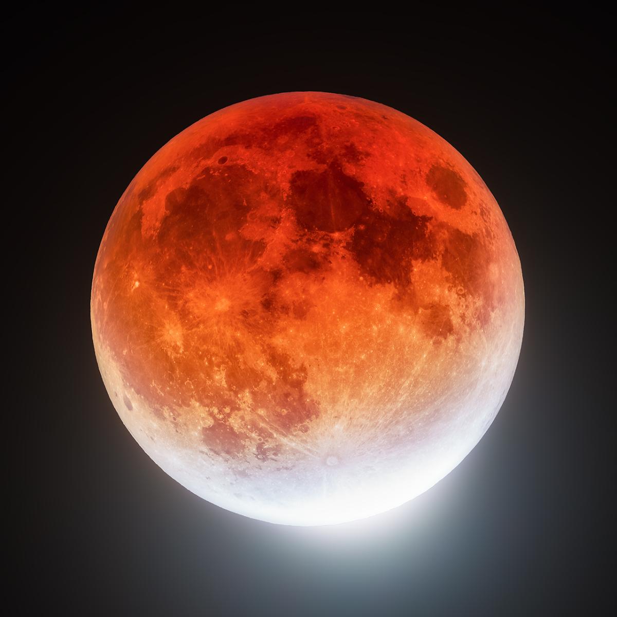 The Moon during an eclipse, lit up a deep orange colour with the white light of the Sun just visible on the bottom right hand side