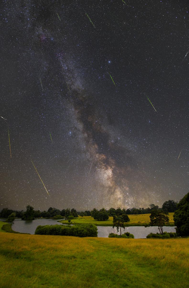 Night skyscape showing the Perseid Meteor Shower and the Milky Way, with stars appearing to cascade down to a scene of green fields and lakes