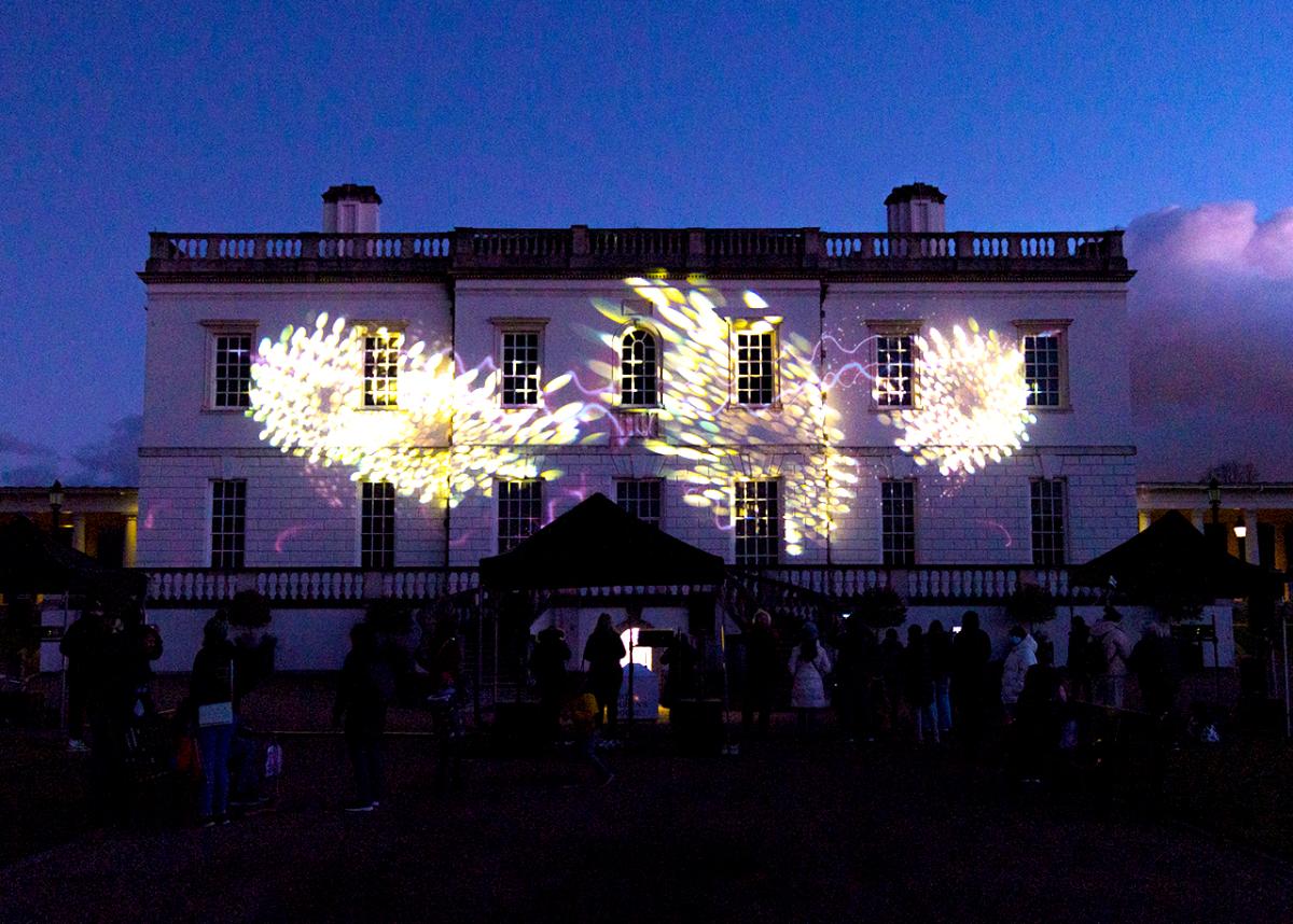 A light projection designed to look like fireworks is beamed onto the façade of the Queen's House at dusk. Bright white and yellow lights can be seen 'exploding' on the building's surface