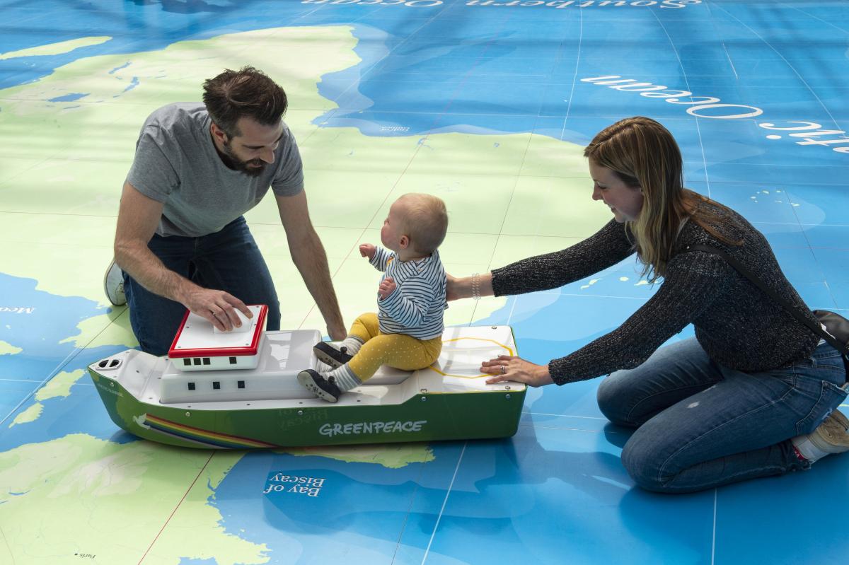 Two adults playing with a toddler who is sitting on a model ship on a big world map printed on the floor