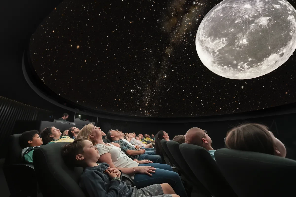 Adults and children reclined in seats, looking up at domed image of sky with big image of the Moon.
