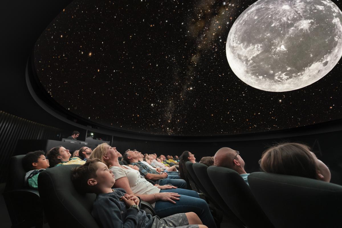 Image from below in the planetarium, showing adults and children reclined in seats looking up at domed image of sky with big image of the Moon