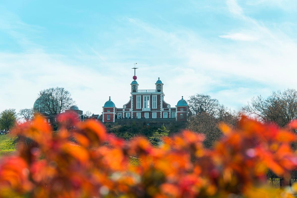 A photograph of the historic Royal Observatory Greenwich. The view is looking up at the brick building from Greenwich Park, with autumnal leaves in the foreground and a clear blue sky above