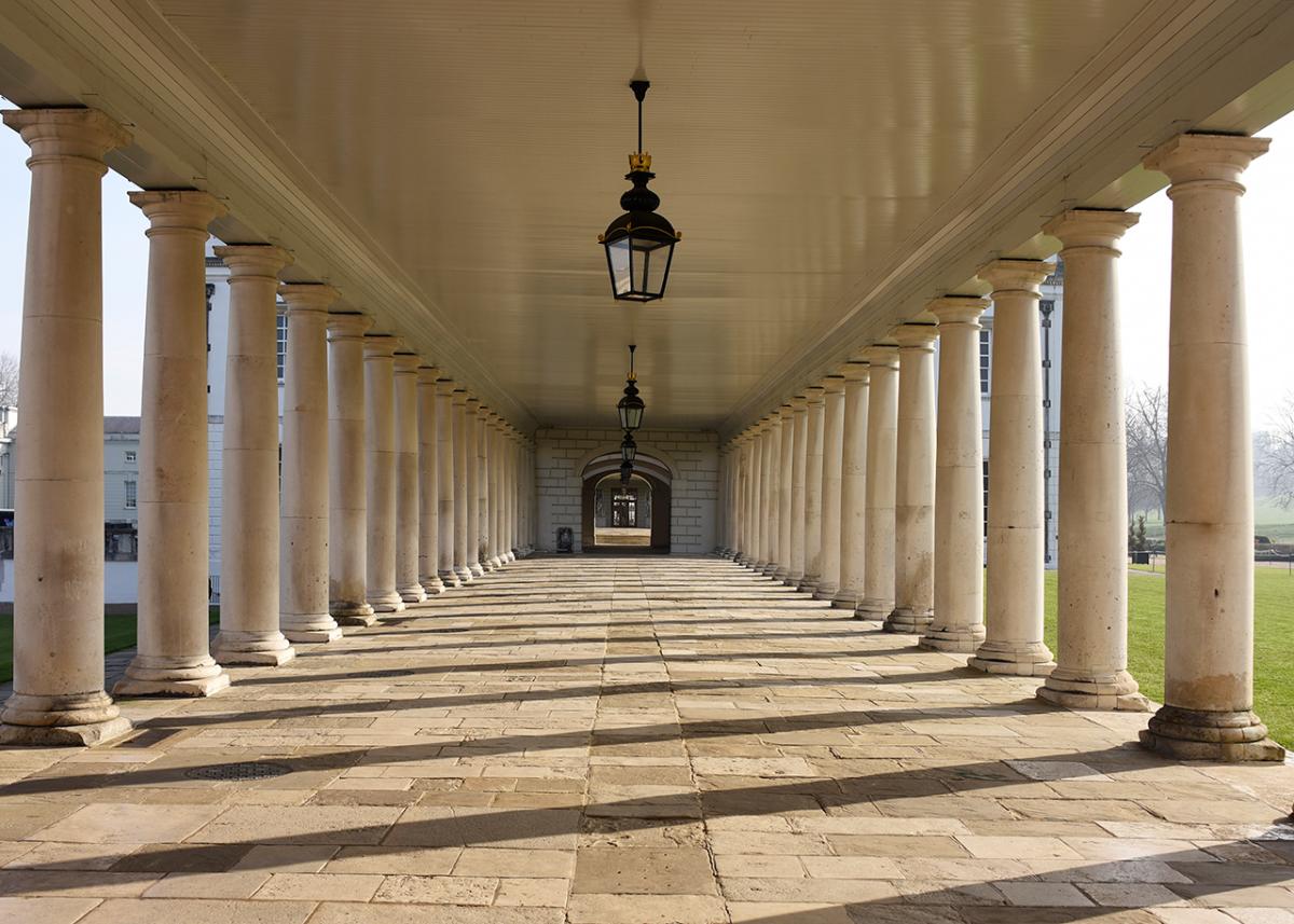 View looking down a stone walkway with grey columns either side. Sunlight is casting shadows across the pavement, adding to the grandeur of the view