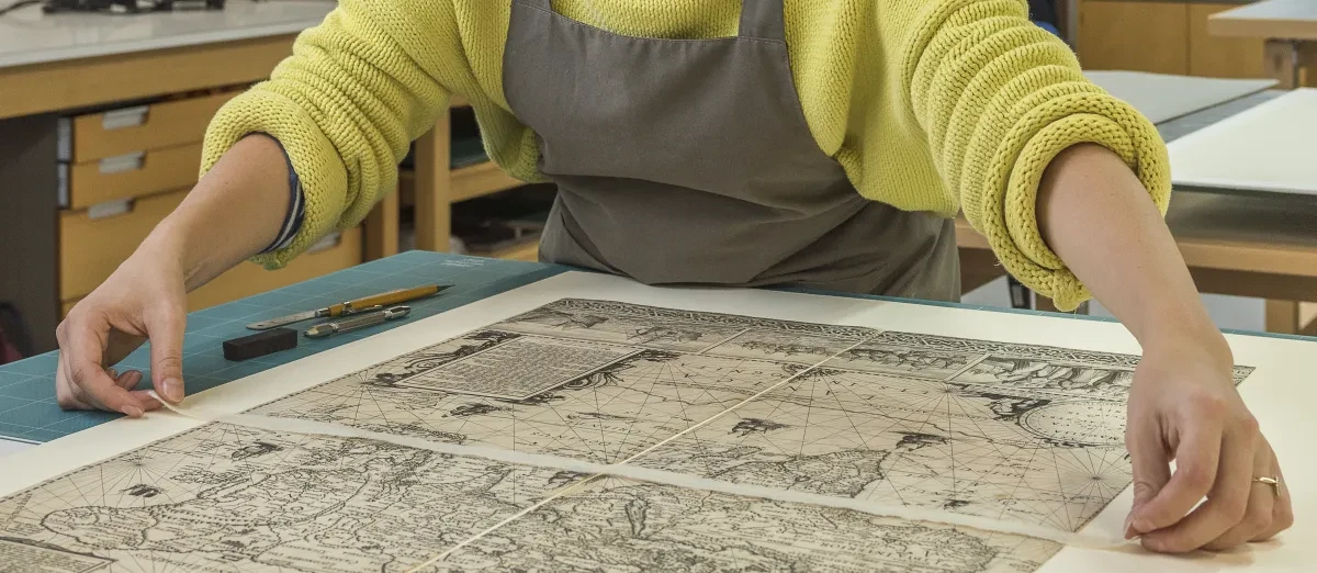 A conservator wearing an apron overlooks a work on paper.