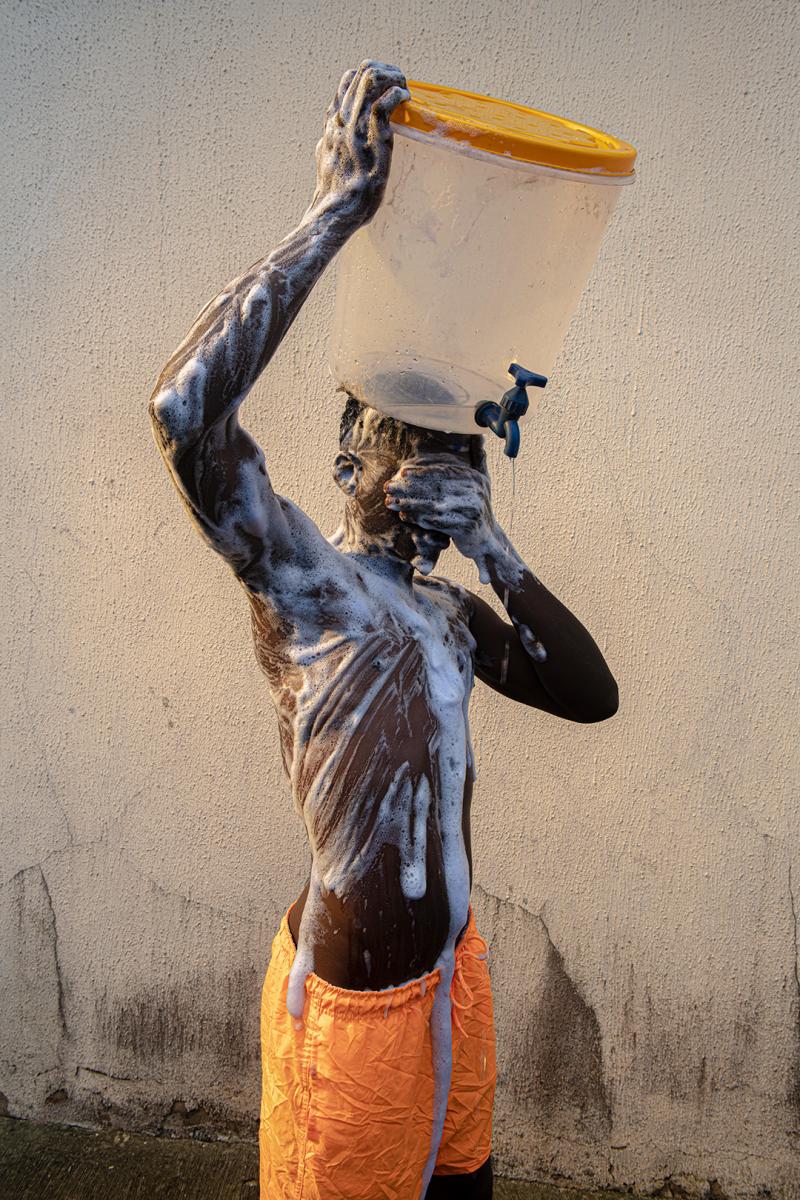 A man in orange swim shorts washes himself in the street. His body is soapy and he is balancing a bucket of water on his head with a tap in the bottom